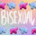 Stegosaurus charms placed in an alternating pattern along two rows on the top and bottom of the image. One charm is magenta with pastel blue back plates and pastel purple eyes. The other is pastel blue with pastel purple plates and magenta eyes. "Bisexual" is handwritten in white in the middle of the image between the two rows. The background is an unevenly faded white grid overlaying a pastel rainbow gradient.
