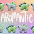 Alternating stegosaurus charms against a pastel rainbow gridded background. One is light green with black back plates and grey eyes. The other is light grey with light green plates and black eyes. They line the word "Aromantic", written in white, on the top and bottom of the image.